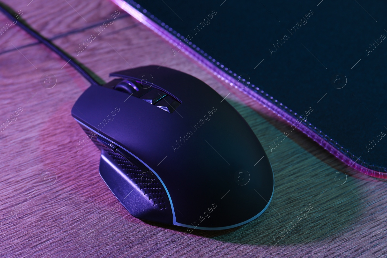 Photo of Black computer mouse and mousepad on wooden table in neon lights, closeup