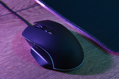 Photo of Black computer mouse and mousepad on wooden table in neon lights, closeup