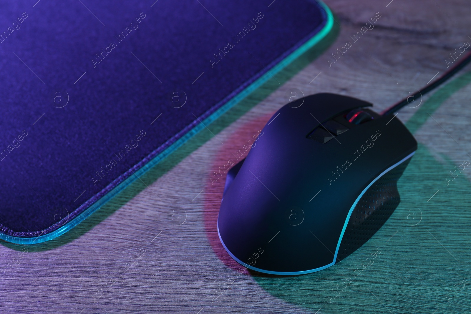 Photo of Black computer mouse and mousepad on wooden table in neon lights, closeup