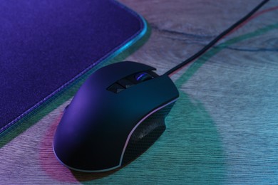 Photo of Black computer mouse and mousepad on wooden table in neon lights, closeup