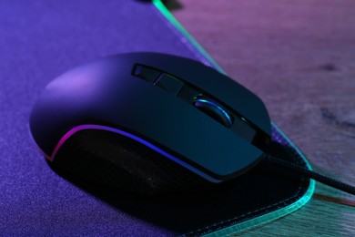 Photo of Black computer mouse and mousepad on wooden table in neon lights, closeup