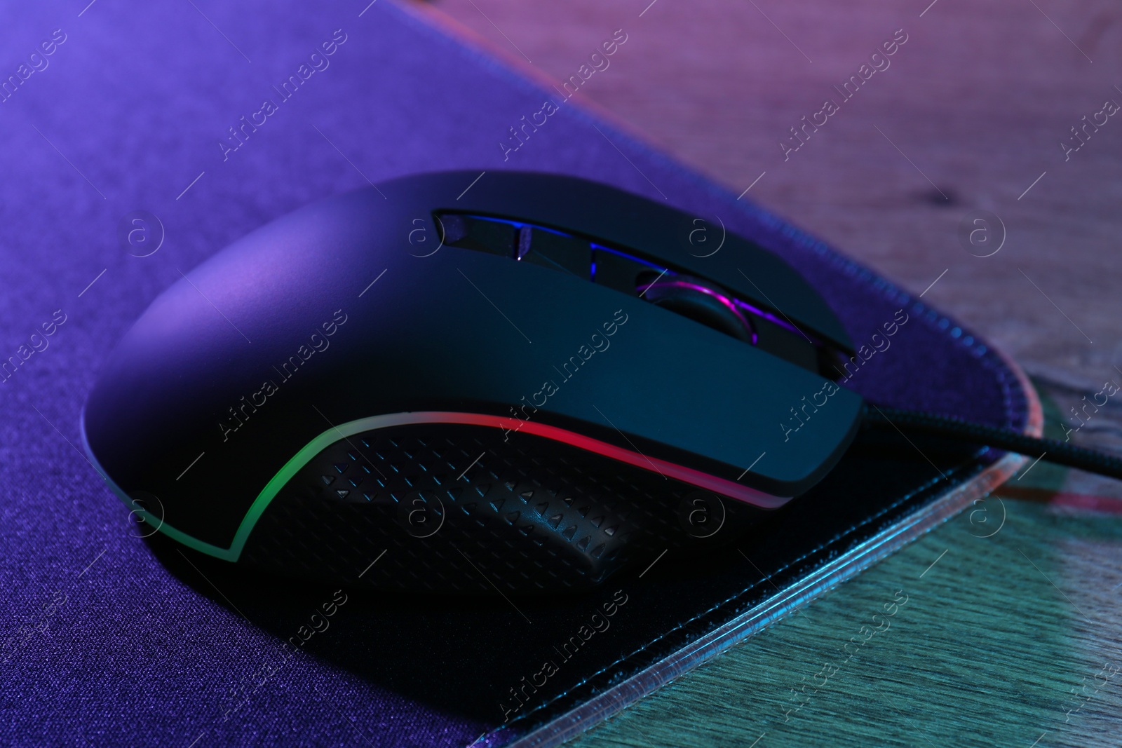 Photo of Black computer mouse and mousepad on wooden table in neon lights, closeup