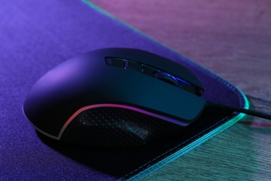 Photo of Black computer mouse and mousepad on wooden table in neon lights, closeup