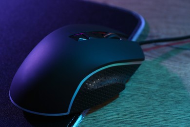 Photo of Black computer mouse and mousepad on wooden table in neon lights, closeup