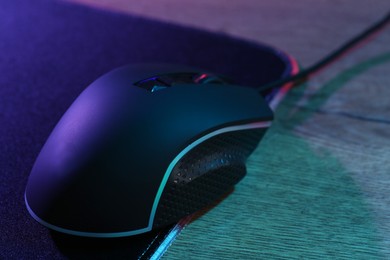 Photo of Black computer mouse and mousepad on wooden table in neon lights, closeup