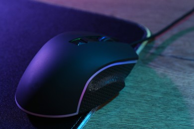 Photo of Black computer mouse and mousepad on wooden table in neon lights, closeup