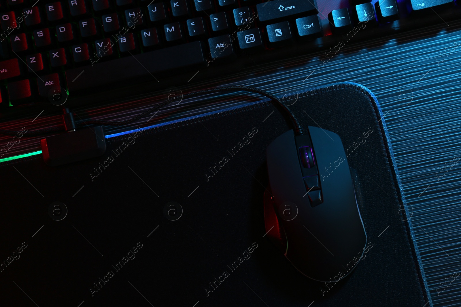 Photo of Computer mouse, mousepad and keyboard on dark wooden table in neon lights, flat lay