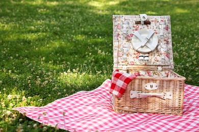 Photo of One picnic wicker basket with checkered napkin, tableware and blanket on green grass. Space for text