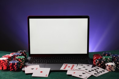 Photo of Laptop, poker chips and playing cards on green table. Online game