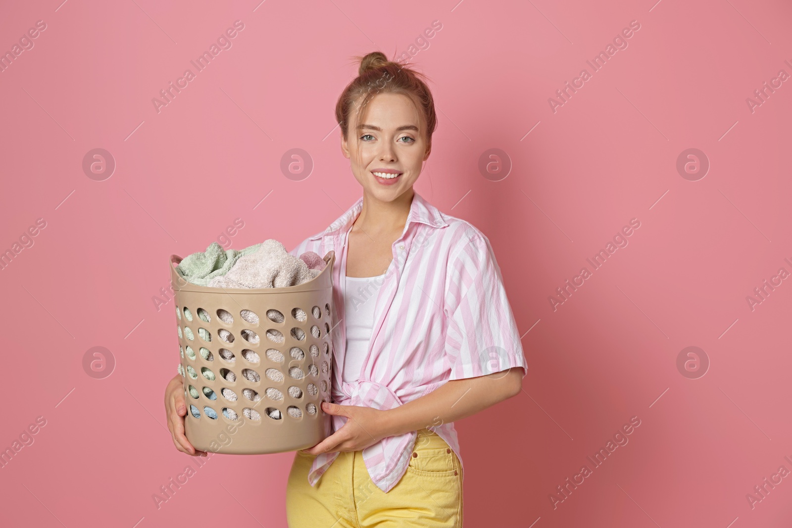 Photo of Happy young housewife with basket full of laundry on pale pink background