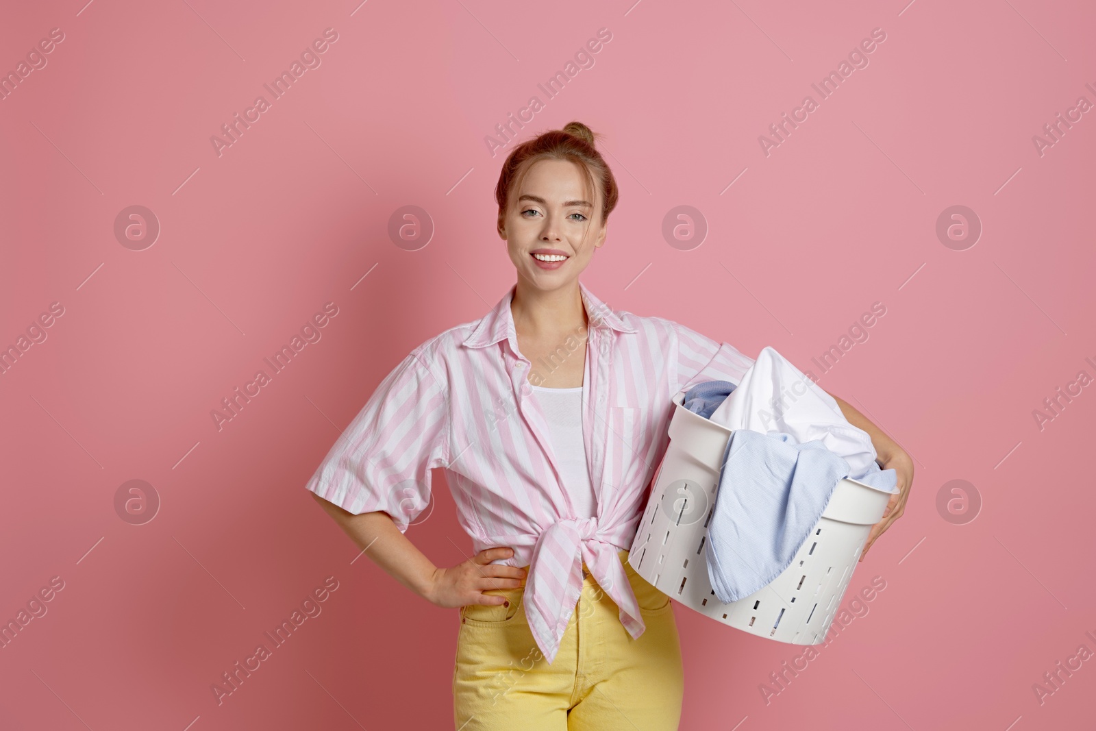 Photo of Happy young housewife with basket full of laundry on pale pink background