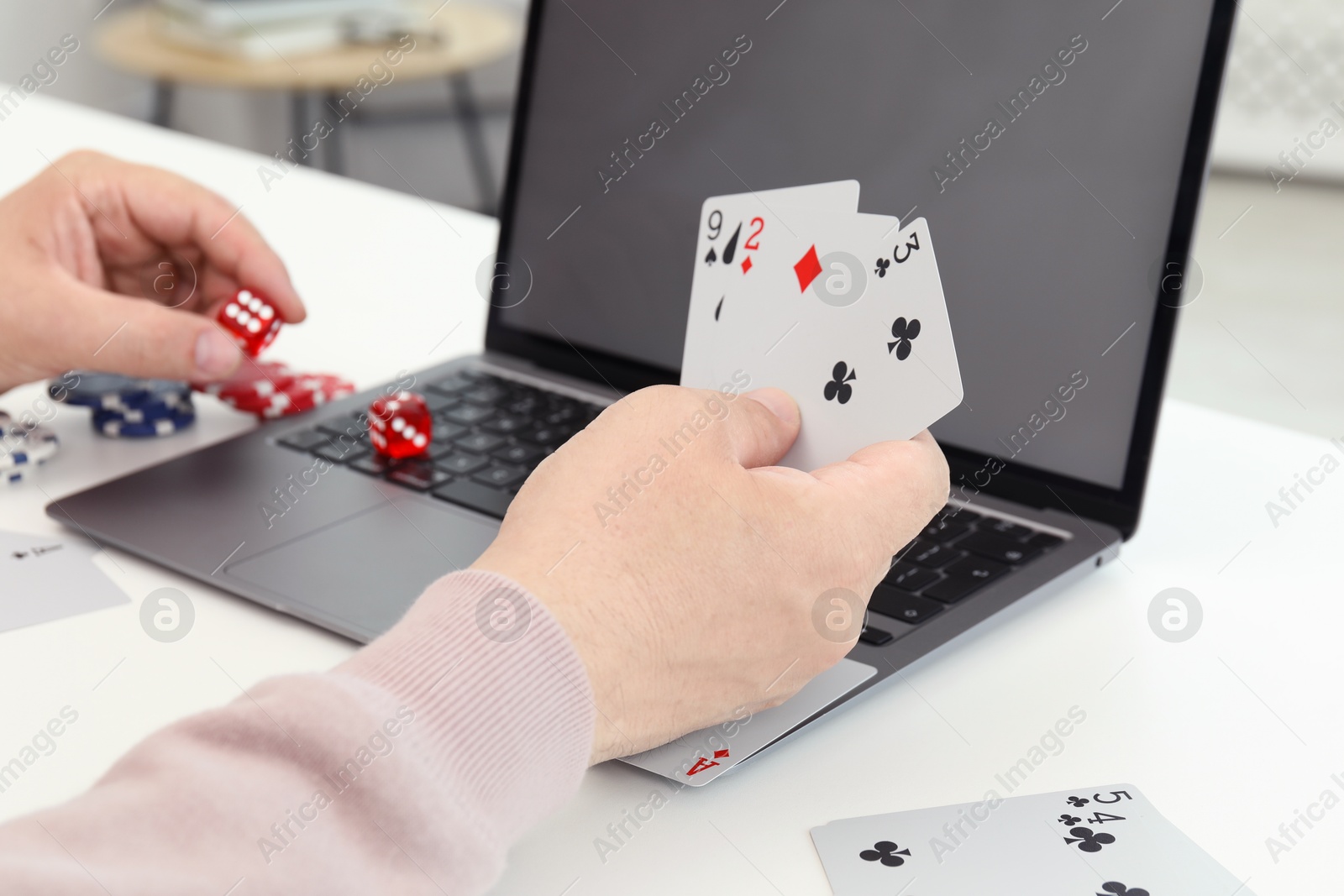 Photo of Online poker. Man holding playing cards and dice at white table with laptop, closeup