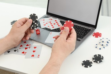 Photo of Online poker. Man holding chips at white table with laptop, playing cards and dice, closeup