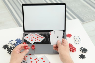 Photo of Online poker. Man holding playing cards and dice at white table with laptop, closeup