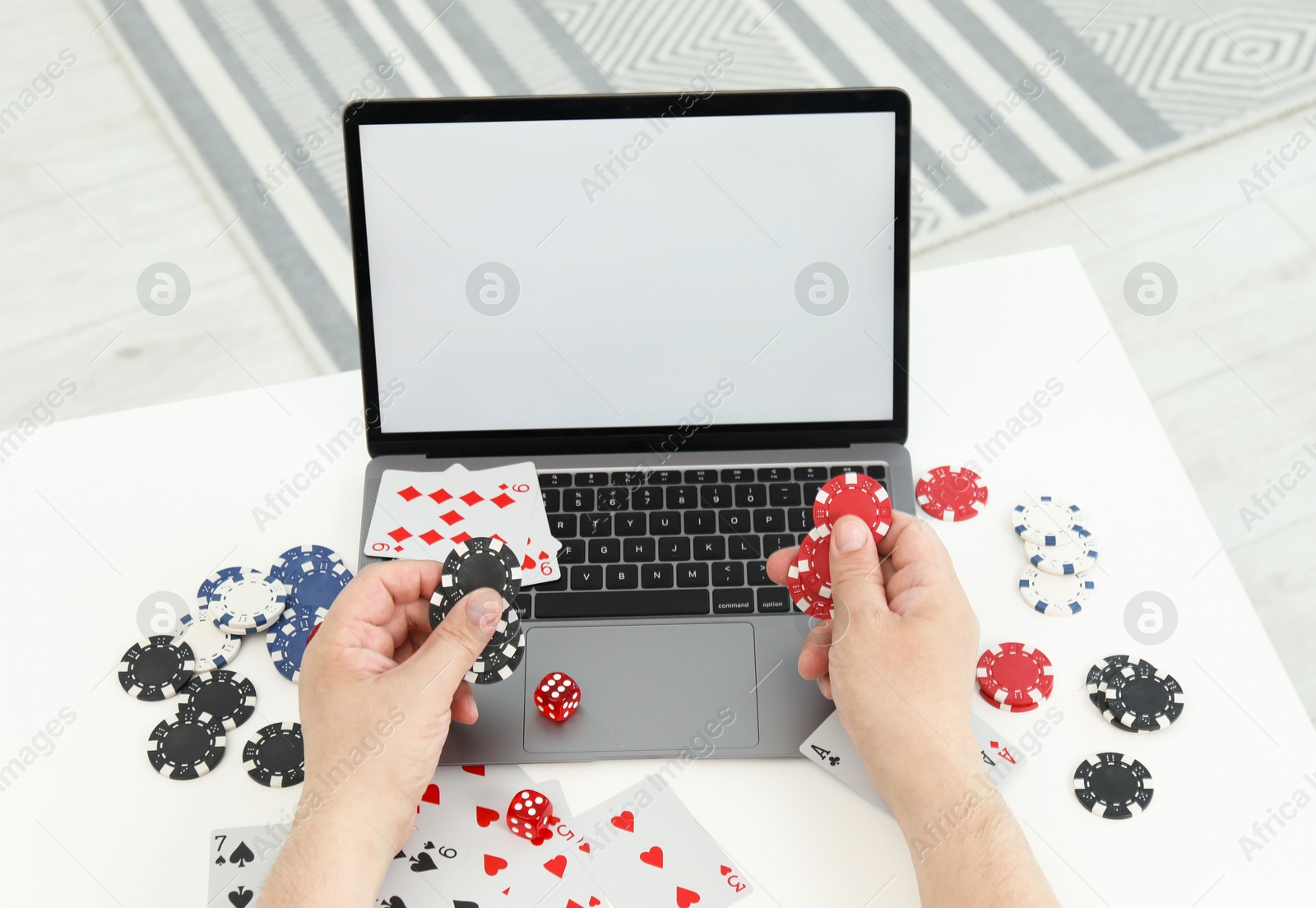 Photo of Online poker. Man holding chips at white table with laptop, playing cards and dice, closeup