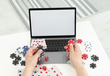 Photo of Online poker. Man holding chips at white table with laptop, playing cards and dice, closeup