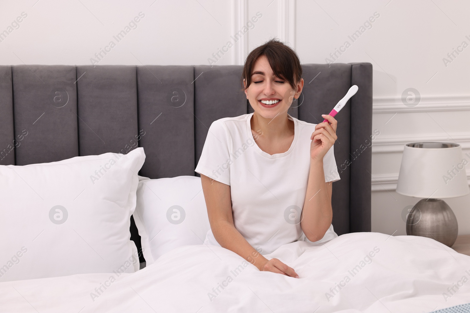 Photo of Happy young woman with pregnancy test on bed at home