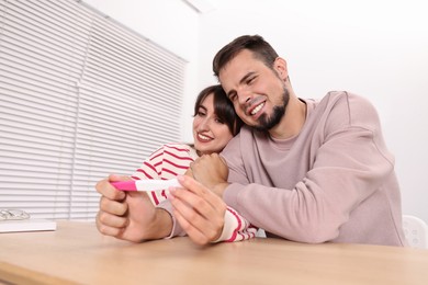 Photo of Happy young couple with pregnancy test at table indoors, wide angle lens effect