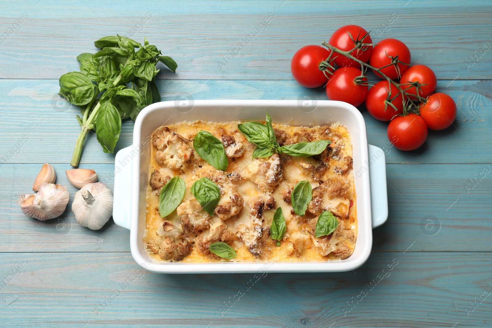 Photo of Tasty baked cauliflower in baking dish, basil, tomatoes and garlic on light blue wooden table, top view