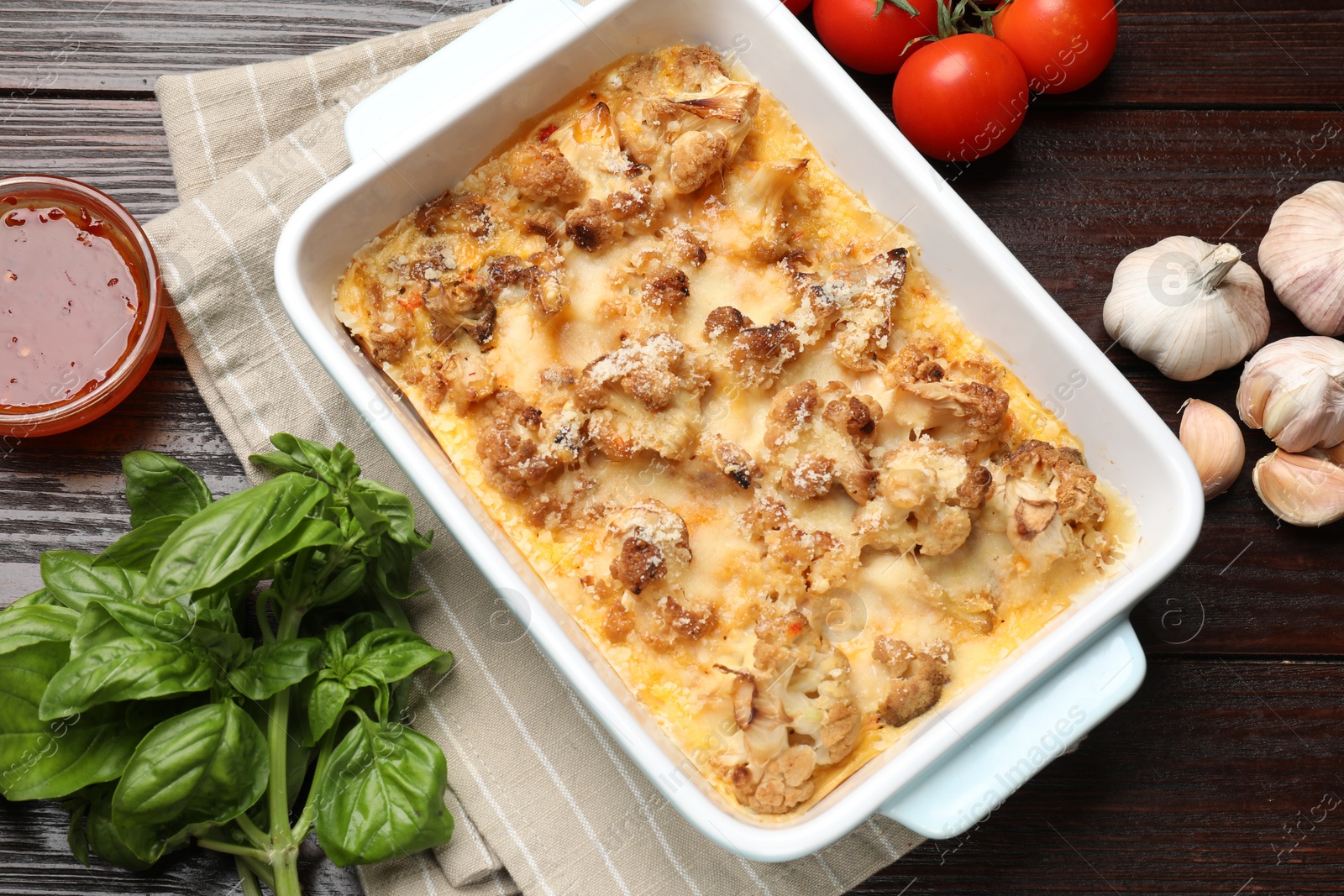 Photo of Tasty baked cauliflower in baking dish, basil, tomatoes, sauce and garlic on wooden table, top view