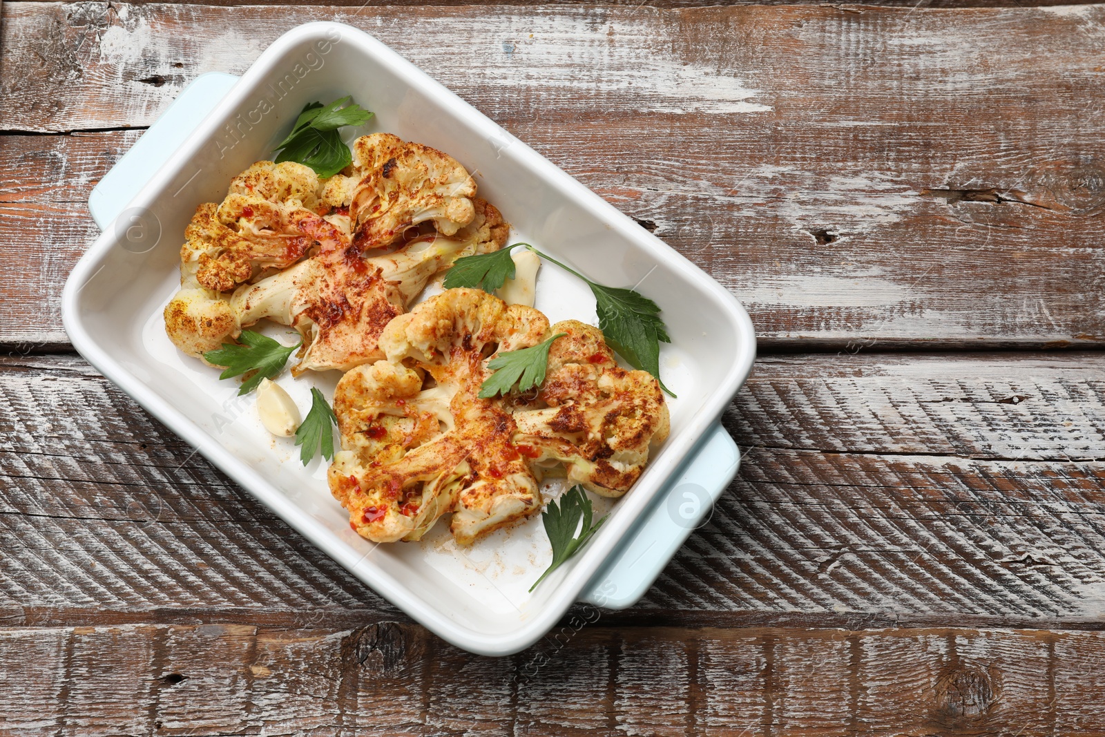 Photo of Tasty cauliflower steaks, parsley and garlic in baking dish on wooden table, top view. Space for text