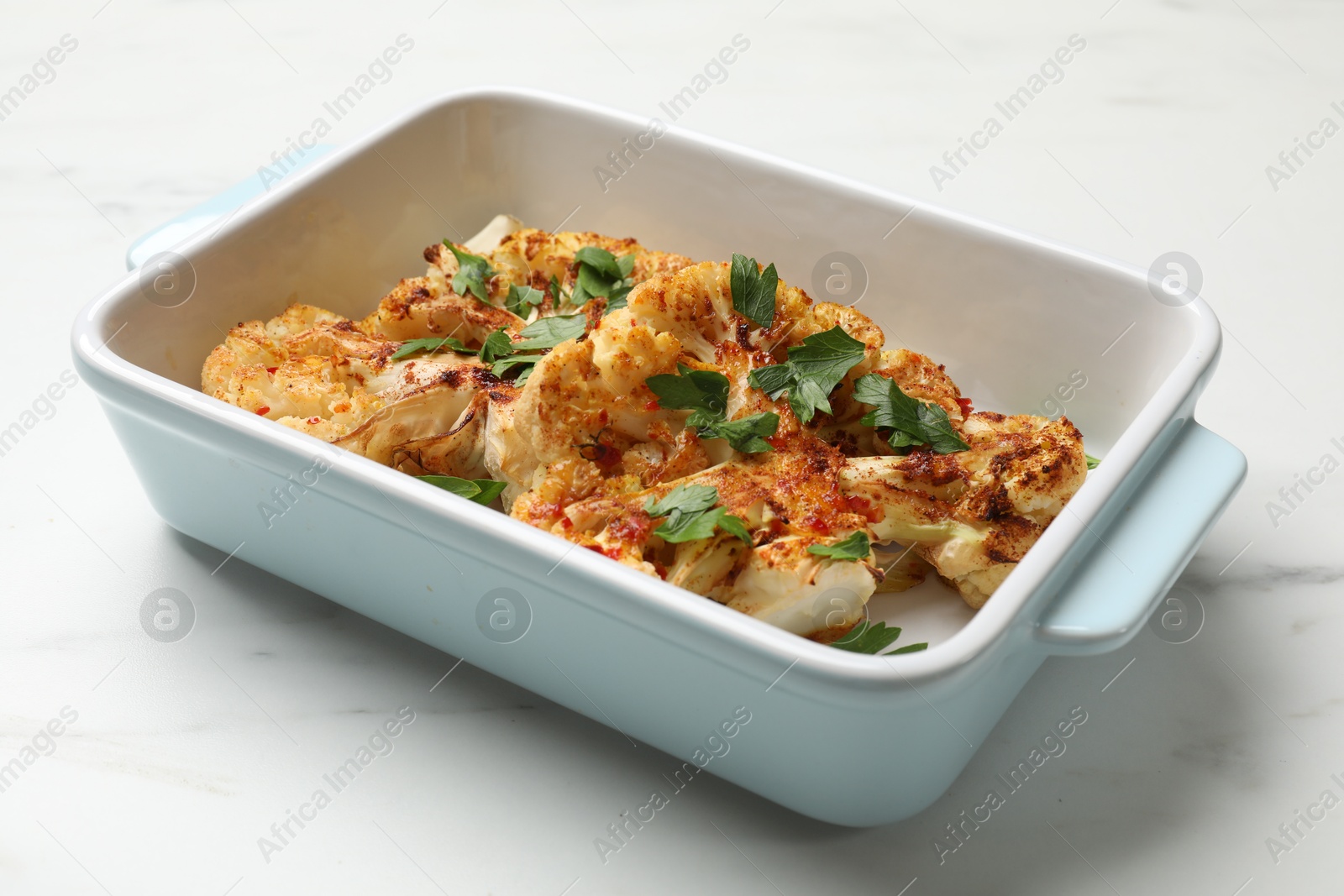 Photo of Tasty cauliflower steaks and parsley in baking dish on white marble table, closeup