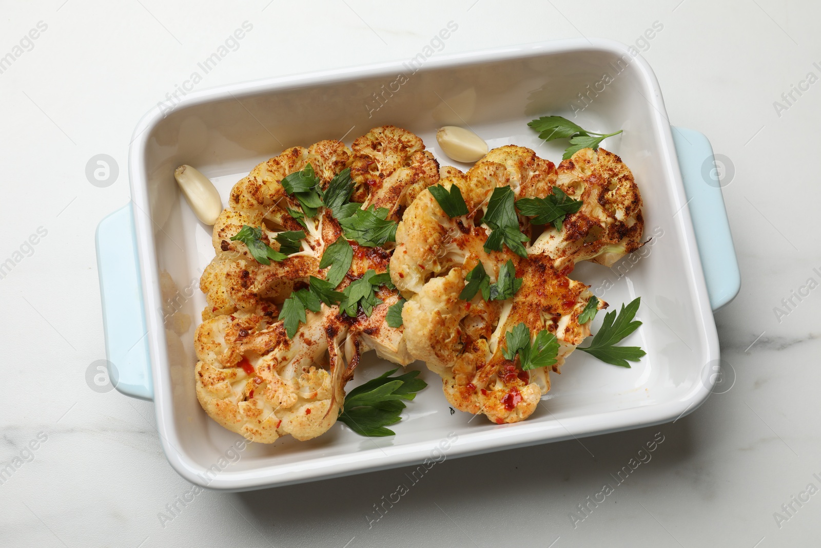 Photo of Tasty cauliflower steaks and parsley in baking dish on white marble table, top view