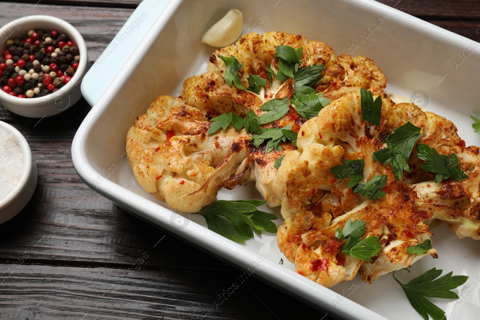 Photo of Tasty cauliflower steaks in baking dish and spices on wooden table, closeup