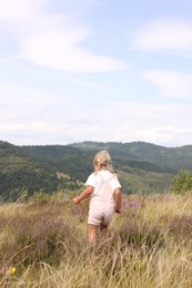 Cute little girl standing on meadow near mountains, back view