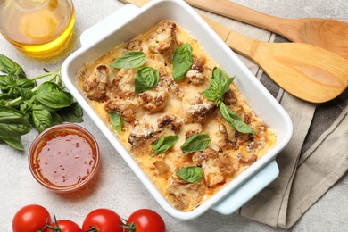 Tasty baked cauliflower in baking dish, basil, sauce, tomatoes and oil on light grey table, top view