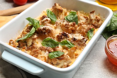 Photo of Tasty baked cauliflower in baking dish, basil and sauce on light grey table, closeup