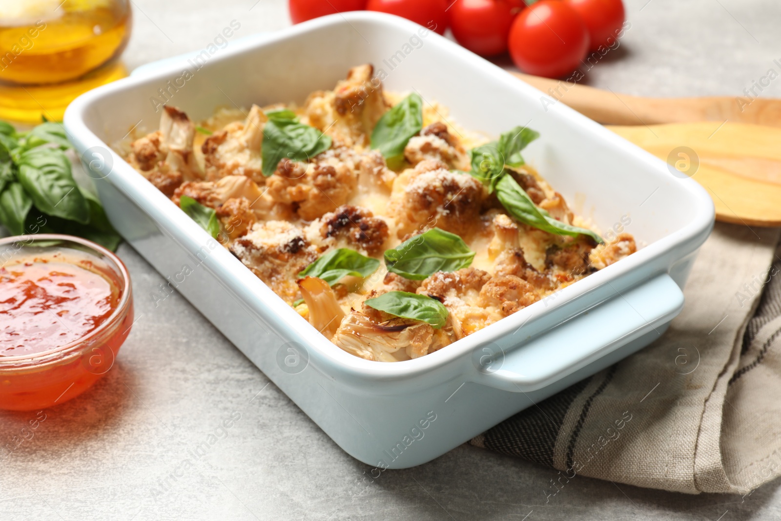 Photo of Tasty baked cauliflower in baking dish, basil and sauce on light grey table, closeup