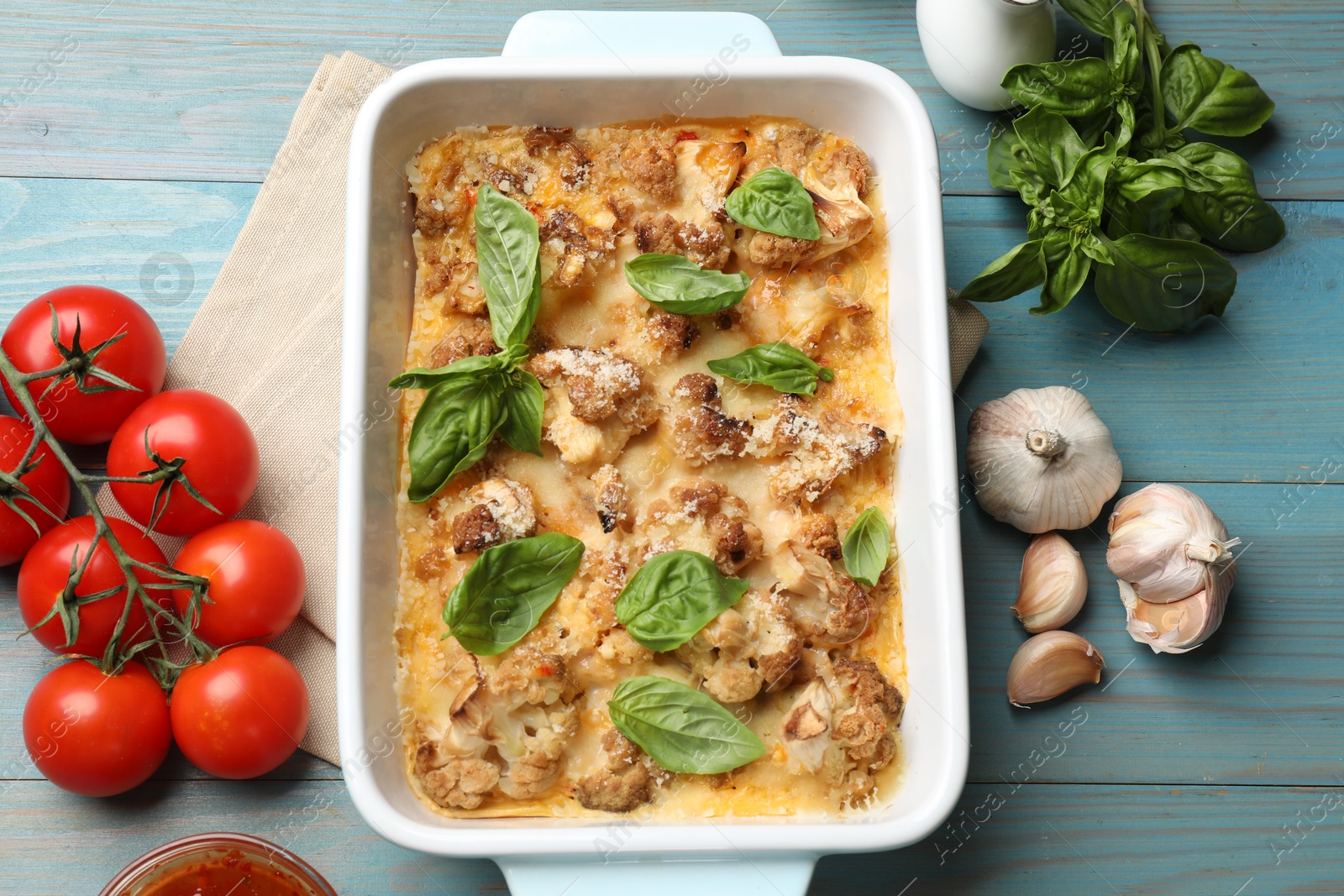 Photo of Tasty baked cauliflower in baking dish, basil, garlic and tomatoes on light blue wooden table, top view