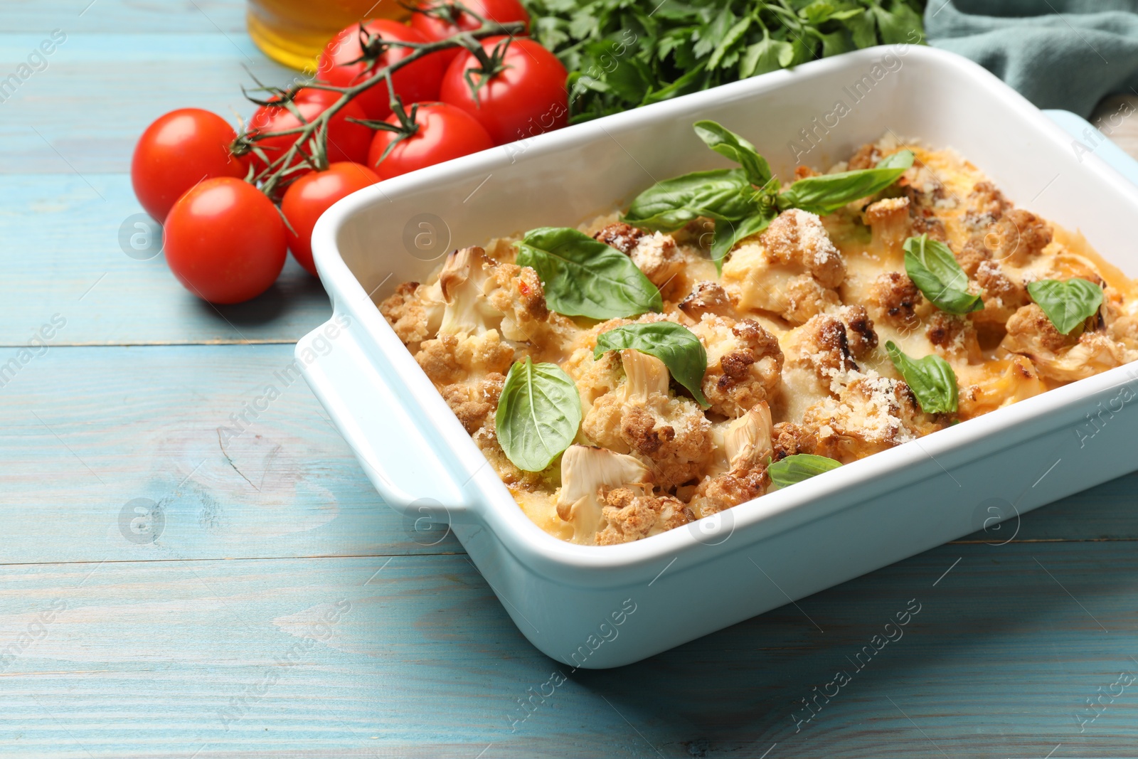 Photo of Tasty baked cauliflower in baking dish, basil, parsley and tomatoes on light blue wooden table, closeup