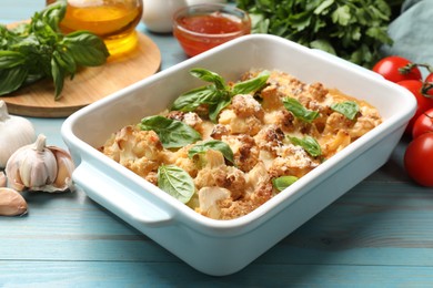 Photo of Tasty baked cauliflower in baking dish, basil, garlic, tomatoes and sauce on light blue wooden table, closeup