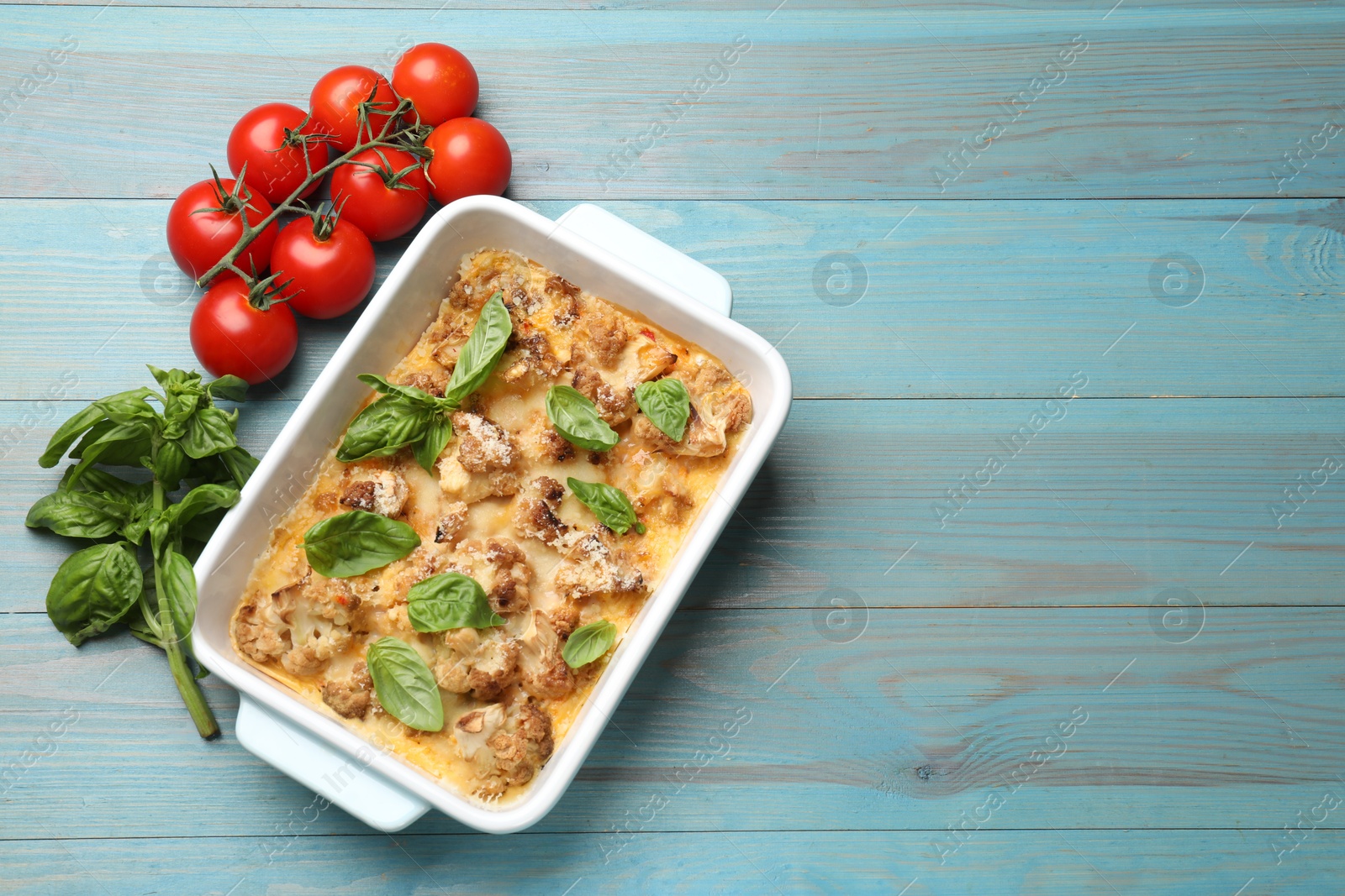 Photo of Tasty baked cauliflower in baking dish, basil and tomatoes on light blue wooden table, top view. Space for text