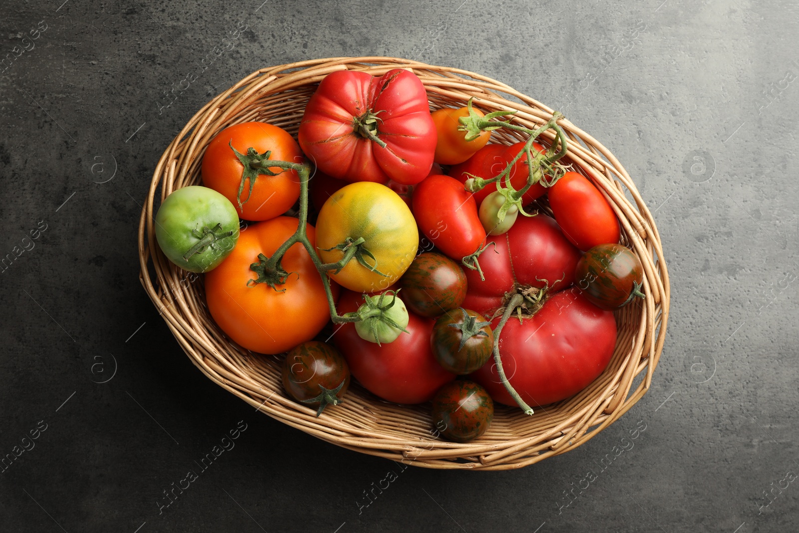 Photo of Different ripe and unripe tomatoes in wicker basket on grey table, top view