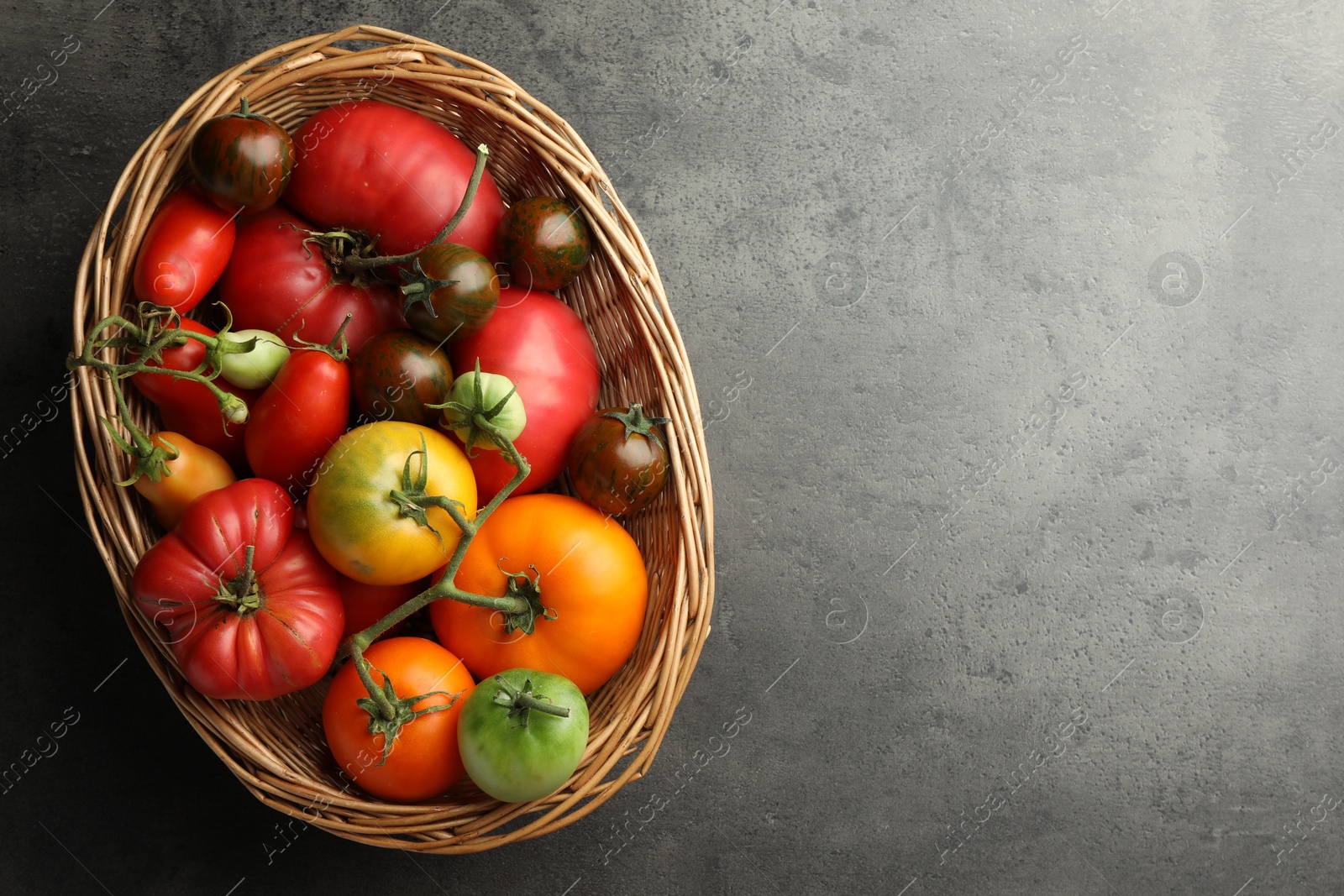 Photo of Different ripe and unripe tomatoes in wicker basket on grey table, top view. Space for text