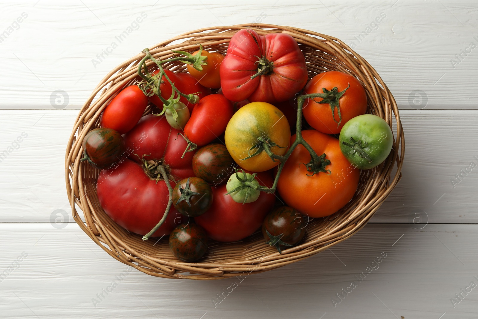 Photo of Different ripe and unripe tomatoes in wicker basket on white wooden table, top view