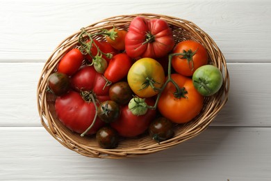 Different ripe and unripe tomatoes in wicker basket on white wooden table, top view