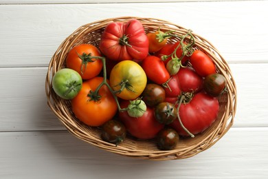 Photo of Different ripe and unripe tomatoes in wicker basket on white wooden table, top view