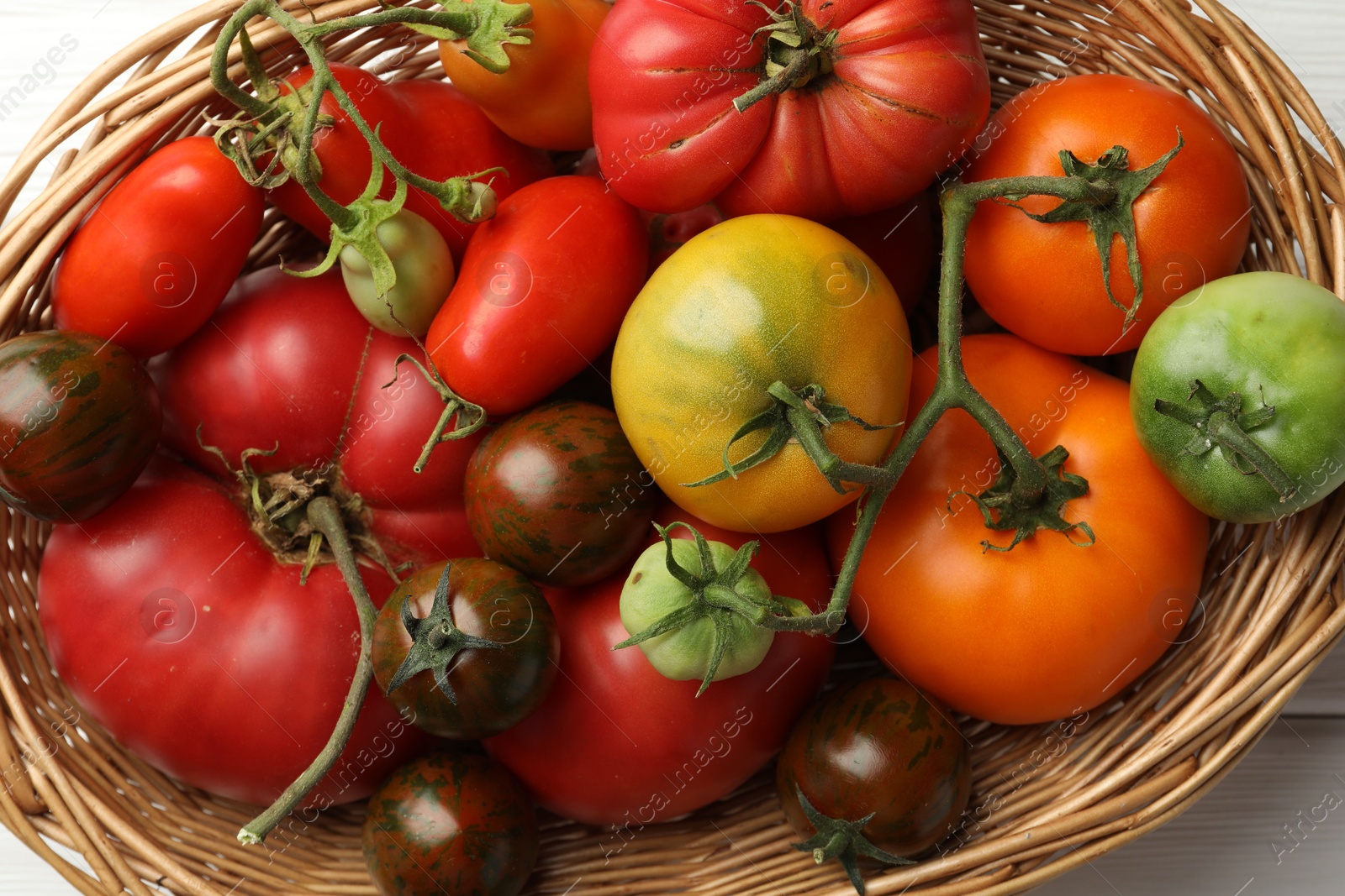 Photo of Different ripe and unripe tomatoes in wicker basket on white wooden table, flat lay