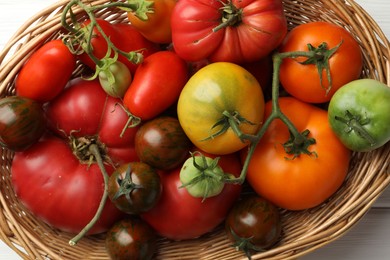 Different ripe and unripe tomatoes in wicker basket on white wooden table, flat lay