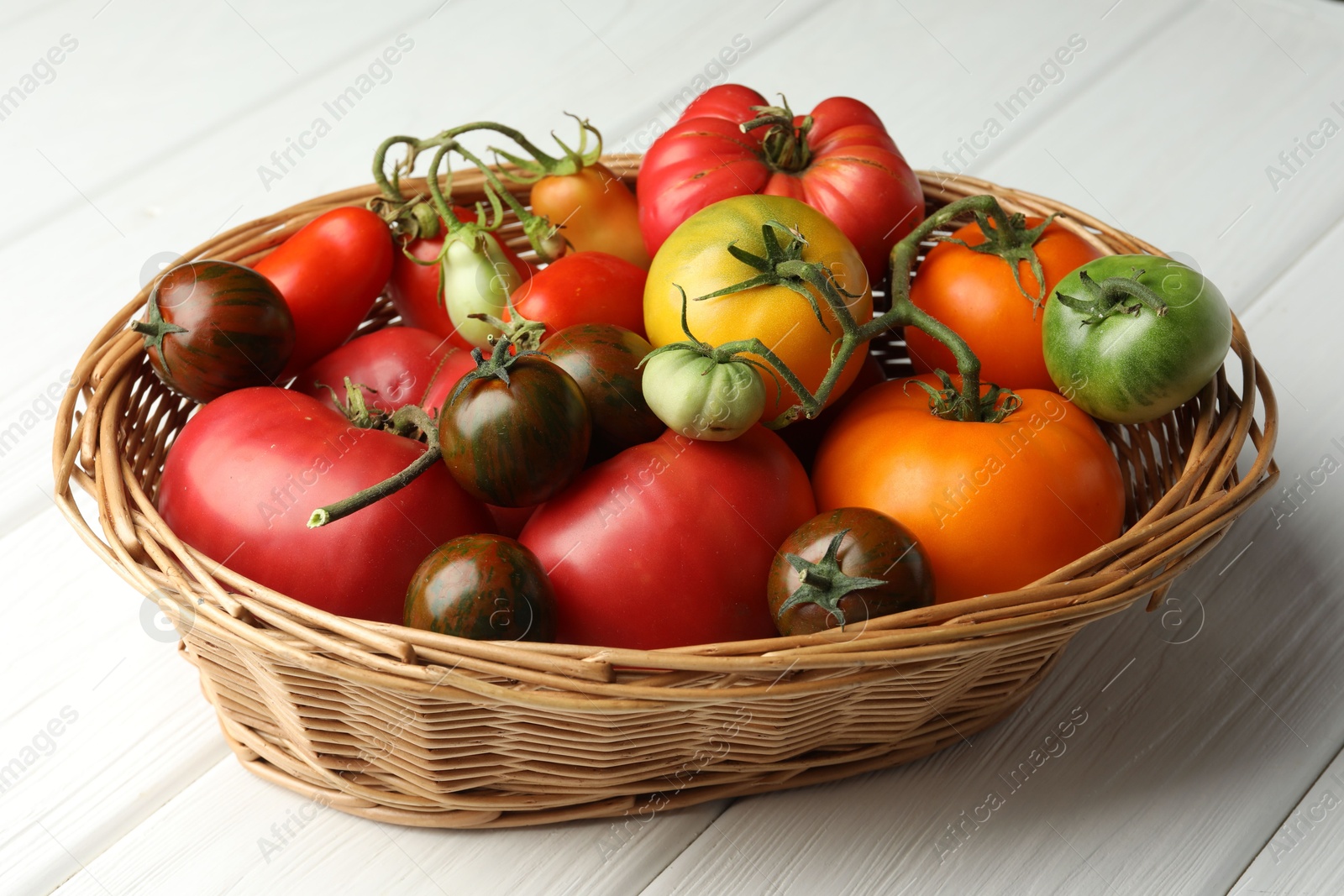 Photo of Different ripe and unripe tomatoes in wicker basket on white wooden table