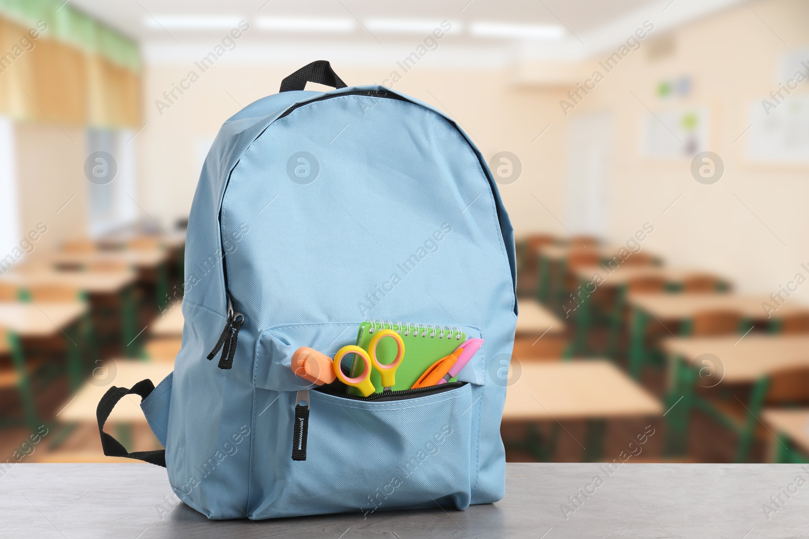 Image of Light blue backpack with stationery on school desk in classroom. Space for text