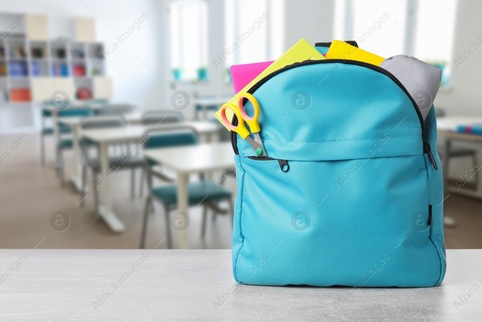 Image of Light blue backpack with stationery on school desk in classroom. Space for text