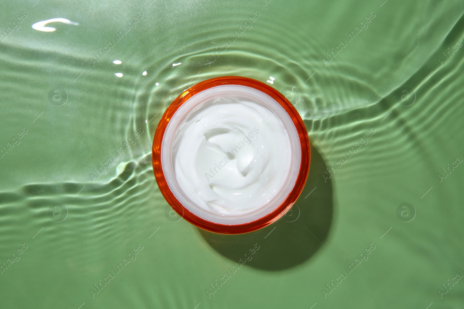 Photo of Jar with cream in water on light green background, top view. Cosmetic product