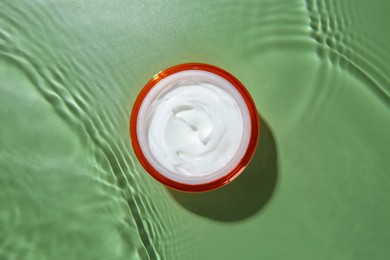 Photo of Jar with cream in water on light green background, top view. Cosmetic product