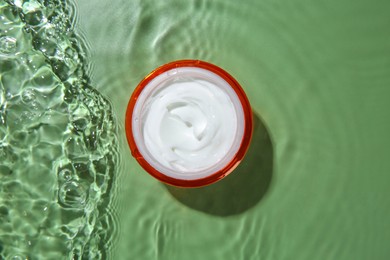 Photo of Jar with cream in water on light green background, top view. Cosmetic product