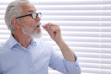 Senior man in glasses taking pill at home, space for text